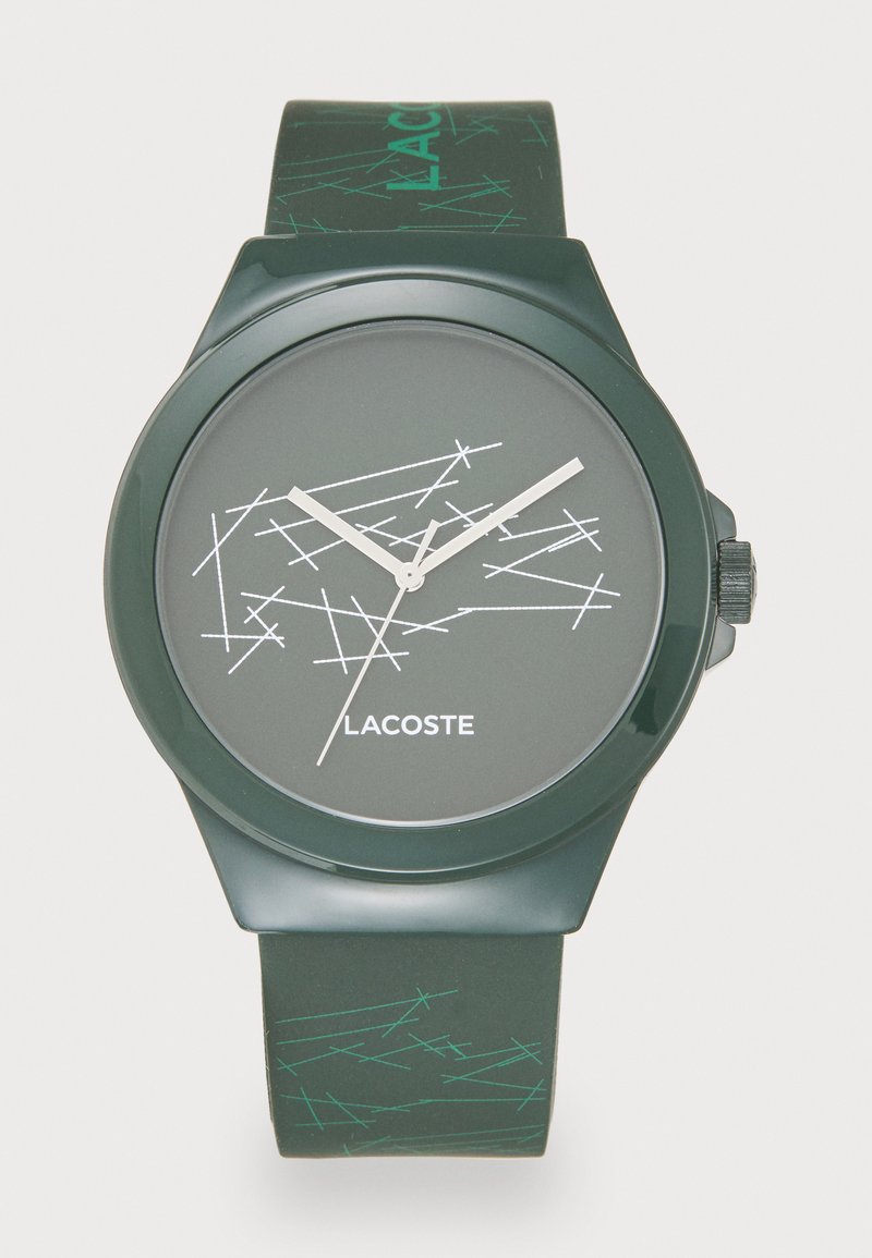 Roheline silikoonkell ümmarguse korpusega, mustal tahvel on valged abstraktsed jooned, hõbedased osutid ja logo. Rihmal on geomeetriline muster.