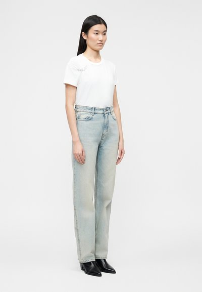 Γυναίκα που φοράει άσπρο κοντομάνικο μπλουζάκι με μικρή λεπτομέρεια φτερού και ανοιχτό μπλε τζιν ψηλόμεσο κανονική γραμμή με μαύρες μυτερές μπότες.