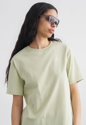 Kvinde med langt mørkt hår iført oversized lysegrøn Jordan t-shirt og store mørke wraparound solbriller, kigger til siden.