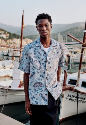 Mladý muž v krátkom vzorovanom tričku stojí pri kotviacich loďkách s horami v pozadí pri západe slnka.