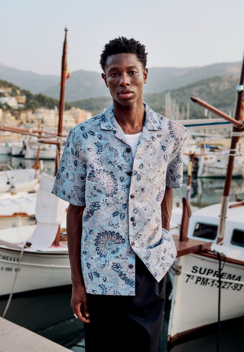 Jeune homme portant une chemise à manches courtes à motifs se tient près de bateaux amarrés avec des montagnes en arrière-plan au coucher du soleil.