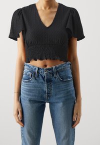 Černý crop top s krátkými nabíranými rukávy, strukturovanou tkaninou, výstřihem do V a volánkovým lemem. Nosený s modrými džíny s vysokým pasem.