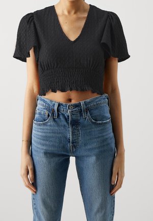 Top corto negro con mangas cortas abullonadas, tela texturizada, escote en V y dobladillo con volantes. Llevado con jeans azul de cintura alta.