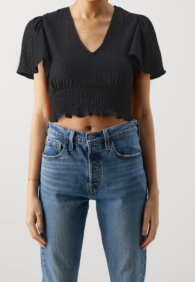 Černý crop top s krátkými nabíranými rukávy, strukturovanou tkaninou, výstřihem do V a volánkovým lemem. Nosený s modrými džíny s vysokým pasem.