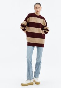 Czerwono-beżowy sweter w paski z dzianiny z okrągłym dekoltem, w oversize'owym fasonie i prążkowaną teksturą, zestawiony z jasnoniebieskimi dżinsami typu straight-leg i masywnymi białymi botkami.