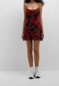 Robe noire avec un motif floral rouge, décolleté froncé et fines bretelles. Portée avec des chaussures de sport blanches et des chaussettes courtes sur un fond clair.