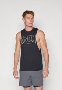 Fekete ujjatlan trikó, amelyen egy kiemelkedő fehér "IRON" grafika látható elöl. Szürke sportnadrággal viselve. Puha pamut anyagból készült.