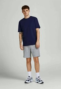 Jovem vestido com uma camiseta azul marinho, calções cinzentos, meias brancas e ténis de desporto, posando contra um fundo claro e neutro.