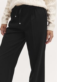 Schwarz weite Hose mit Kordelzugbund, mit zwei seitlichen Taschen und glatter Textur. Kombiniert mit einem cremefarbenen Sweater aus Kabelausschnitt.