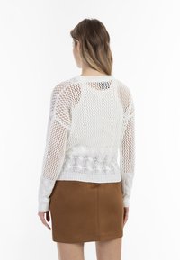 Bílý pletený svetr s síťovinovým vzorem, ribbed manžetami a texturovaným tělem. Kombinován s hnědými sukněmi, ukazuje zadní pohled.