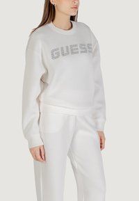 Biała bluza z ribbowanymi mankietami i dołem, z logo "GUESS" wysadzanym srebrnymi dżetami na przodzie, w komplecie z pasującymi białymi dresami.