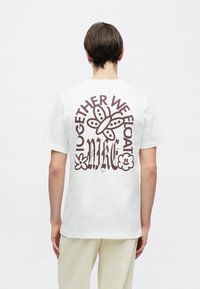 Biała bawełniana koszulka z krótkimi rękawami, z brązowym napisem "TOGETHER WE FLOAT" i ilustracją motyla na plecach.