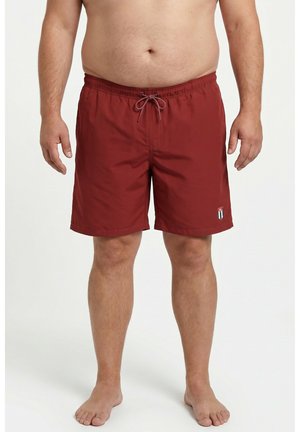 Homme portant un short de bain bordeaux longueur genou avec cordon et petit logo sur la jambe gauche, debout pieds nus sur fond blanc.