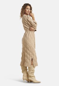 Abito beige lavorato a maglia con motivo a diamante, maniche lunghe e spacco frontale. Abbinato a stivali beige morbidi al ginocchio.