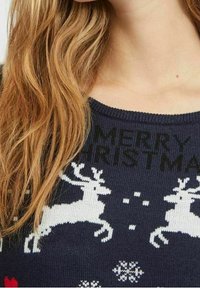 Pull en maille bleu foncé avec des motifs de rennes blancs et des motifs de fête, avec le texte "MERRY CHRISTMAS" affiché en noir de manière bien visible.