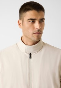 Beige leichte Jacke mit hohem Kragen und Reißverschluss. Glatte Textur und minimalistisches Design ohne zusätzliche Muster oder Akzente.