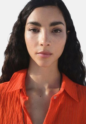 Camicia arancione con colletto in un tessuto texturizzato e stropicciato. La modella ha capelli neri mossi e un'espressione neutra. Lo sfondo è bianco puro.