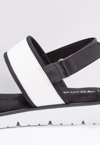 Sandale noir et blanc avec bride noire réglable et bande blanche à l’avant sur une semelle blanche rainurée.