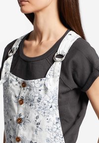 Salopette à motif floral en blanc et bleu marine, ornée de grands boutons en bois et de quincaillerie en métal, portée sur une chemise gris foncé.