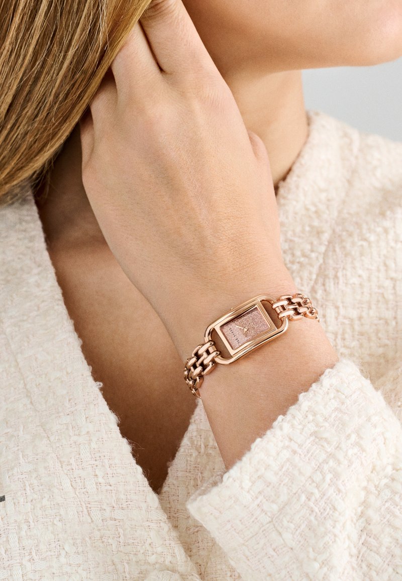 Roosa kuldne metallkäevõru koos ristkülikukujulise kellanäidikuga, millel on tekstureeritud roosakas taust. Ahelakujuline disain koos sileda riistvara aktsendiga.