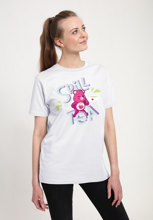 T-shirt en coton blanc avec un graphique coloré d'un ours rose et le texte "Spill the Tea" accompagné d'éclaboussures et d'une tasse de thé. Manches courtes.