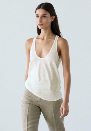 Donna con capelli lunghi e scuri, che indossa una canotta bianca con scollo a U e pantaloni beige, in piedi davanti a uno sfondo chiaro uniforme.