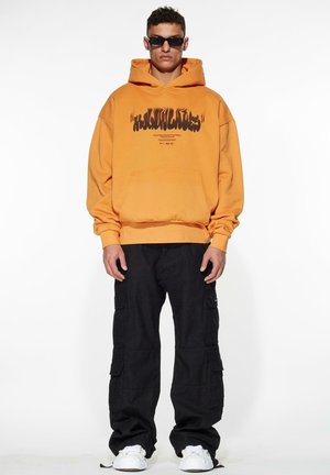Orange bomulds hoodie med sort grafisk tekst, frontlomme og ribbet kant, parret med sorte cargo-bukser og hvide sneakers.