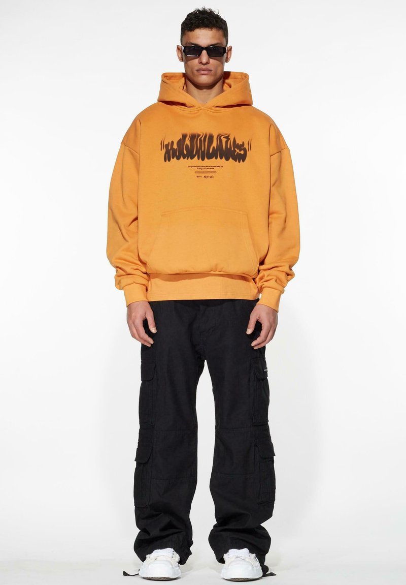 Orange cotton hoodie with black graphic text, front pocket, and ribbed hem, paired with black cargo pants and white sneakers.