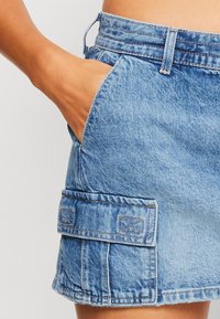 Modré džínové šortky s roztrhaným lemem, vybavené boční kapsou s klopou a švy, a středně vysokým pasem.