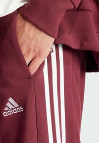 Bordo sporta bikses, kas izgatavotas no kokvilnas sajaukuma, ar trim baltām svītrām sānos un Adidas logotipu priekšpusē. Tekstūra ir gluda.