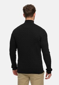 Svart kabelstickad tröja med hög krage, långa ärmar och strukturerat mönster. Figursydd design som följer kroppen, i kontrast till ljusare byxor.