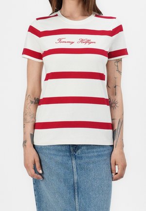 Osoba nosząca białą koszulkę z czerwonymi poziomymi paskami i napisem "Tommy Hilfiger", w połączeniu z niebieską dżinsową spódnicą oraz widocznymi tatuażami na ramionach.