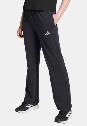 Personne portant un pantalon de survêtement Adidas noir avec le logo sur la cuisse et des baskets Adidas noires à rayures blanches.