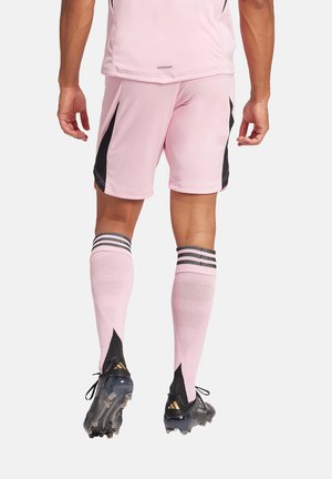 Roze voetbaluniform met zwarte accenten, bestaande uit een kortemouwtop en shorts. Zwart-wit gestreepte kniekousen en zwarte voetbalschoenen.
