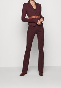 Blazer su misura bordeaux con rever a taglio, chiusura con un bottone e pantaloni aderenti coordinati. Cintura in pelle marrone con fibbia metallica.