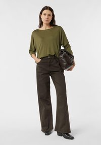 Długi rękaw oliwkowozielonego topu zestawiony z ciemnobrązowymi rozszerzanymi spodniami. Modelka trzyma czarną torebkę. Gładkie faktury o prostych liniach.