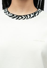 Balti marškinėliai su juodai baltos dryžuotos zebros rašto krašteliu. Ant krūtinės subtilus įspaustas logotipas. Minkštos audinio tekstūros.