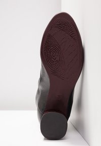 Semelle d'une chaussure en cuir noir avec des motifs de grip texturés et un talon arrondi, positionnée verticalement contre des surfaces blanches.