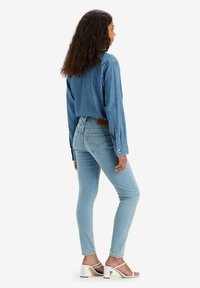 Ljusblå denimjeans med slim fit, kombinerade med en mörk denimknapp-up skjorta. Modellen bär vita blockklacksandaler.
