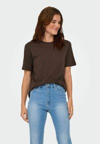 Donna con capelli castani e mossi, che indossa una maglietta a maniche corte marrone scuro infilata in jeans a vita alta blu chiaro, che guarda di lato e sorride.