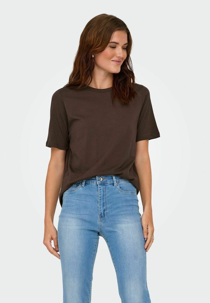 Donna con capelli castani e mossi, che indossa una maglietta a maniche corte marrone scuro infilata in jeans a vita alta blu chiaro, che guarda di lato e sorride.