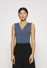 Bodysuit blu senza maniche con scollatura a V profonda, caratterizzato da un design avvolgente e una texture morbida, abbinato a pantaloni neri a vita alta.