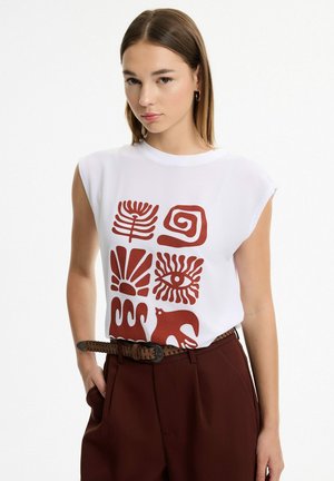Joven mujer con cabello liso que lleva una camiseta blanca sin mangas con símbolos abstractos rojos y pantalones marrones de talle alto.