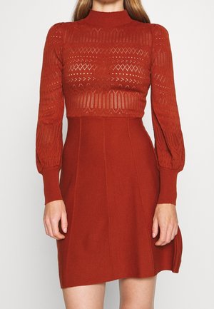 Femme portant une robe tricotée couleur rouille avec un corsage semi-transparent à motif et des manches longues bouffantes, cintrée à la taille et évasée au niveau de la jupe.