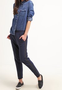 Chemise en denim avec fermeture à boutons et poche poitrine ; associée à des joggers bleu marine avec des rayures latérales et des chaussures à enfiler noires texturées.