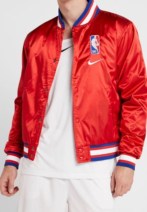 Homme portant une veste NBA en satin rouge avec des poignets et un col rayés bleu, blanc et rouge, par-dessus un t-shirt et un short Nike blancs.