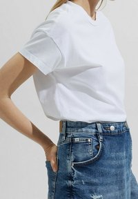 Weißes Baumwoll-T-Shirt mit Rundhalsausschnitt, kurzen Ärmeln und lockerem Schnitt, kombiniert mit einem blauen Jeansrock mit sichtbaren Nähten und Taschen.