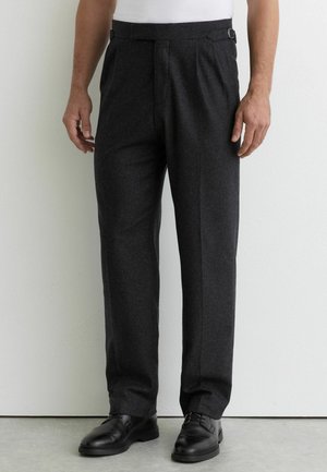 Man draagt donkergrijze op maat gemaakte broek met verstelbare zijbanden, gecombineerd met zwarte nette schoenen, staand tegen een witte muur.