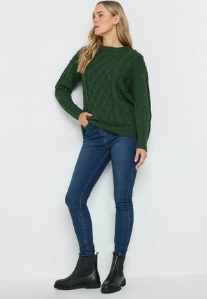 Groene gebreide trui met kabelpatroon, ronde halslijn en schouderknopdetails, gecombineerd met blauwe skinny jeans en zwarte enkellaarzen.