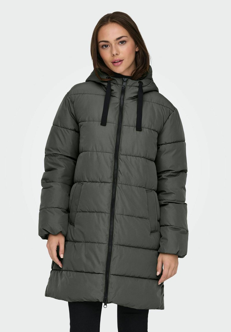 Ung kvinde iført en olivengrøn quilted pufferjakke med hætte og sort lynlås, stående med hænderne afslappet langs siden.
